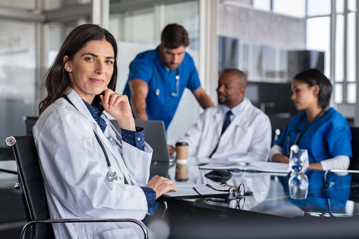 Portrait of a mature, smiling, female doctor sitting in meeting room with specialists and nurses discussing case in background.